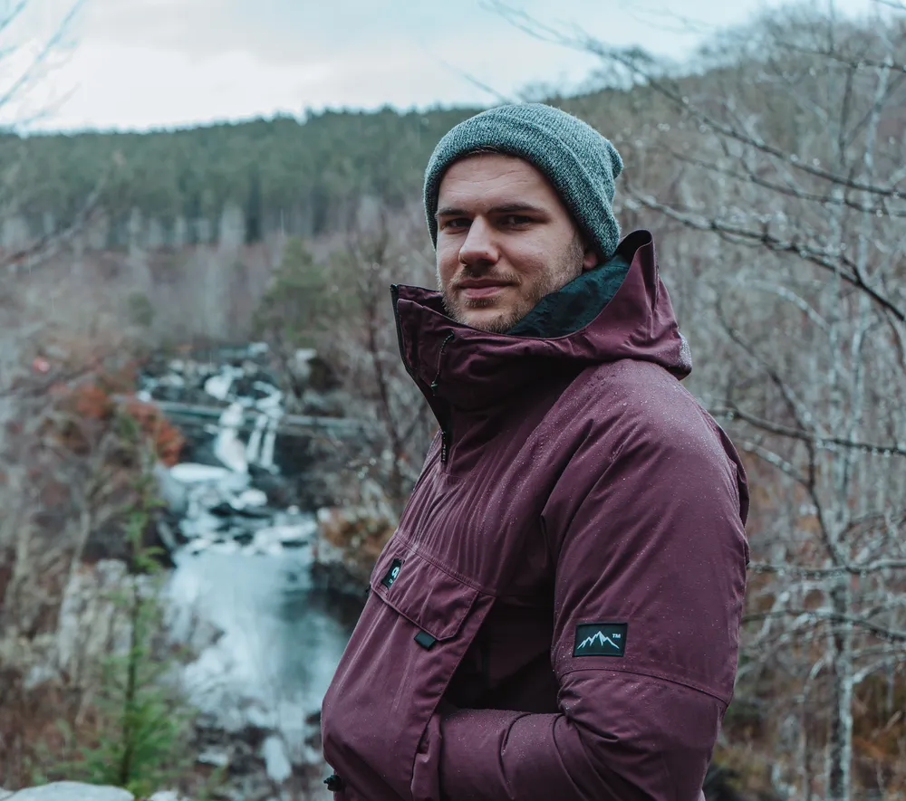 Cam James beside a waterfall, wearing a beanie and jacket
